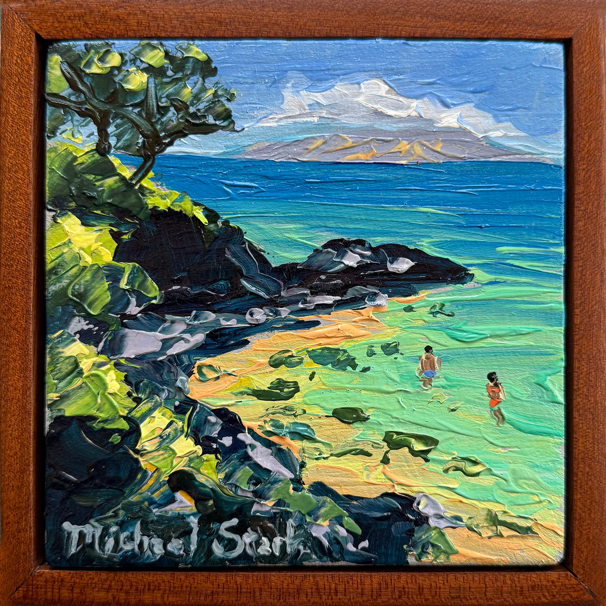 A framed square oil painting depicting a tropical Maui beach scene with turquoise water, dark rocks, green foliage, distant mountains, and two small figures near the shoreline. The painting is signed 'Michael Stark' in the lower left corner and enclosed in a medium brown wooden frame.